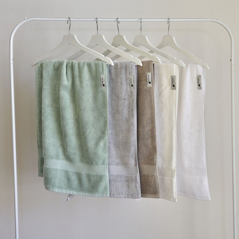 Cinq serviettes en bambou pastel suspendues à des cintres en bois blanc sur fond neutre, affichant vert, gris, marron, crème et