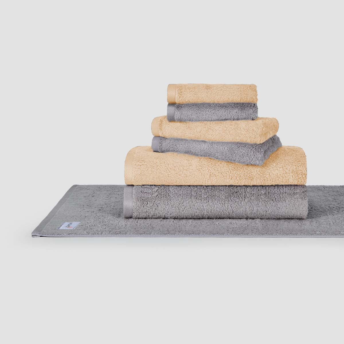 Ensemble de serviettes empilées beige et gris Momo Cotton présenté sur un tapis de bain gris sur fond blanc