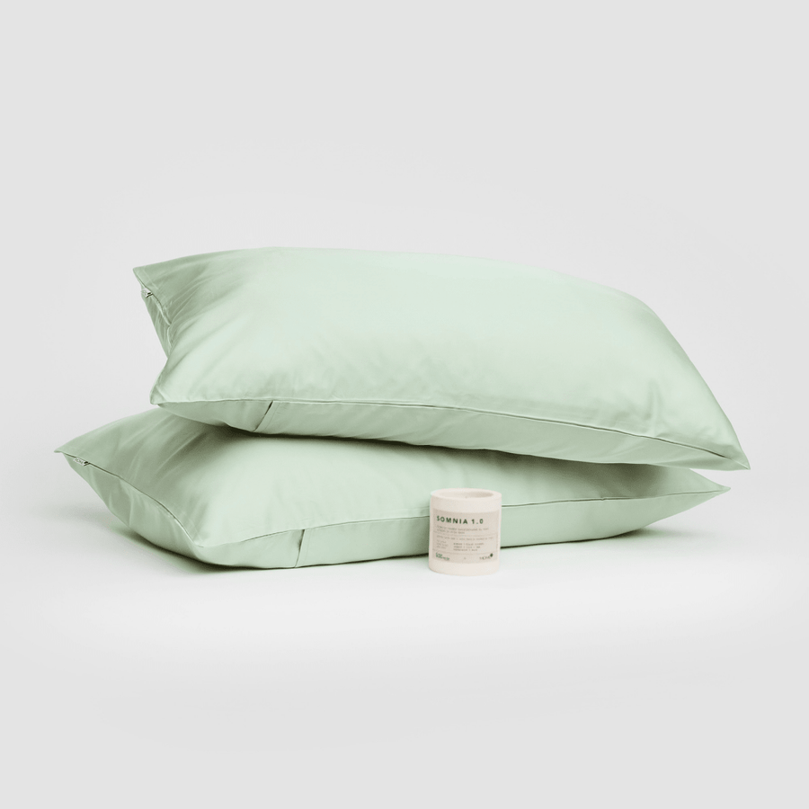 Paire de taies d'oreiller en coton vert sauge empilées avec bougie Somnia 1.0 sur fond blanc