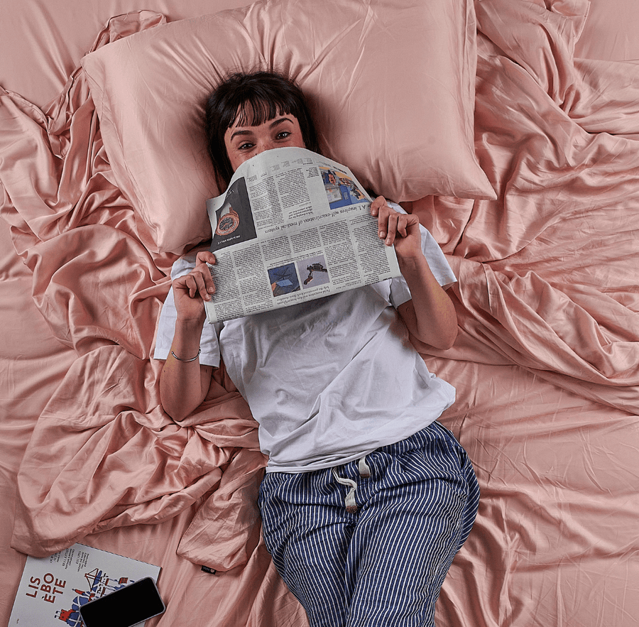 Femme allongée sur un drap de lit en bambou rose lisant un journal, vue de haut
