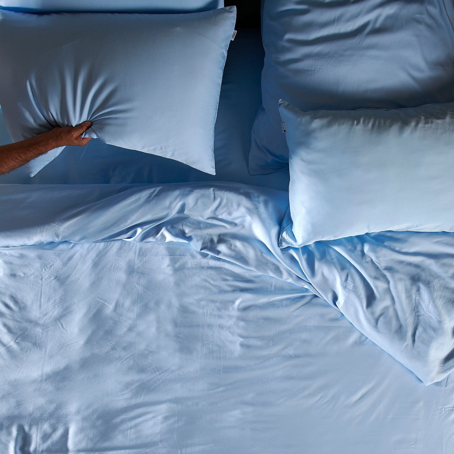 Person arranging light blue bamboo bed sheet set with pillows on bed, showing texture and softness of complete bedding set