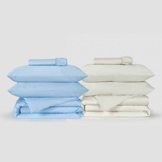 Deux parures de housse de couette Momo Cotton côte à côte, une bleu clair et une crème, avec draps pliés et linge roulé