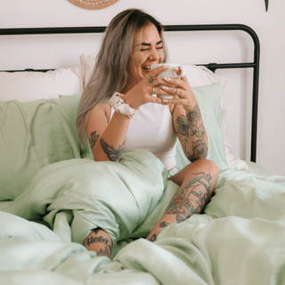 Woman on bed wearing white tank top with sage green bamboo sheet set, displaying soft fabric and comfort